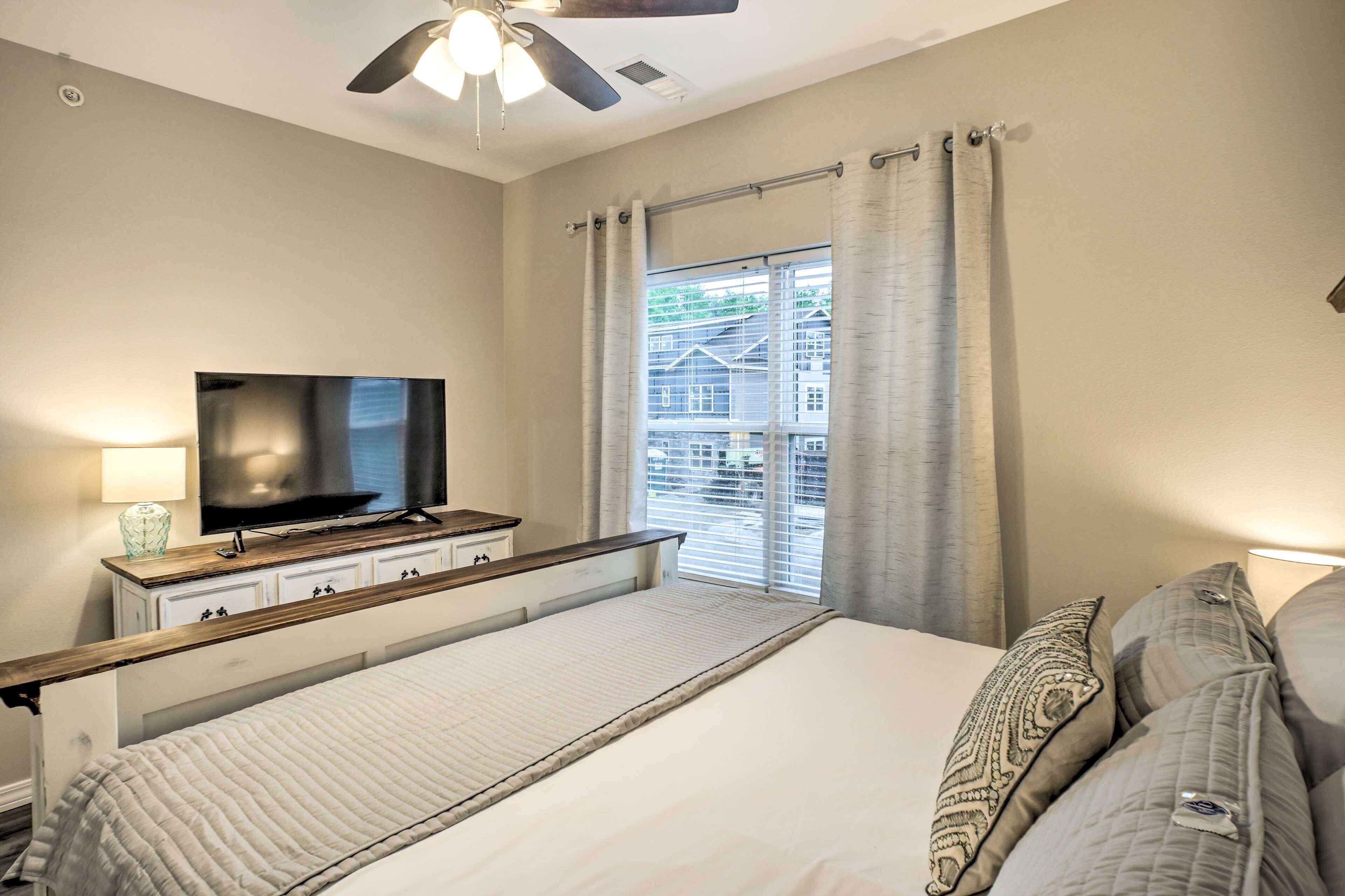A bedroom with a bed, a TV on a dresser, a ceiling fan, and a window with curtains.
