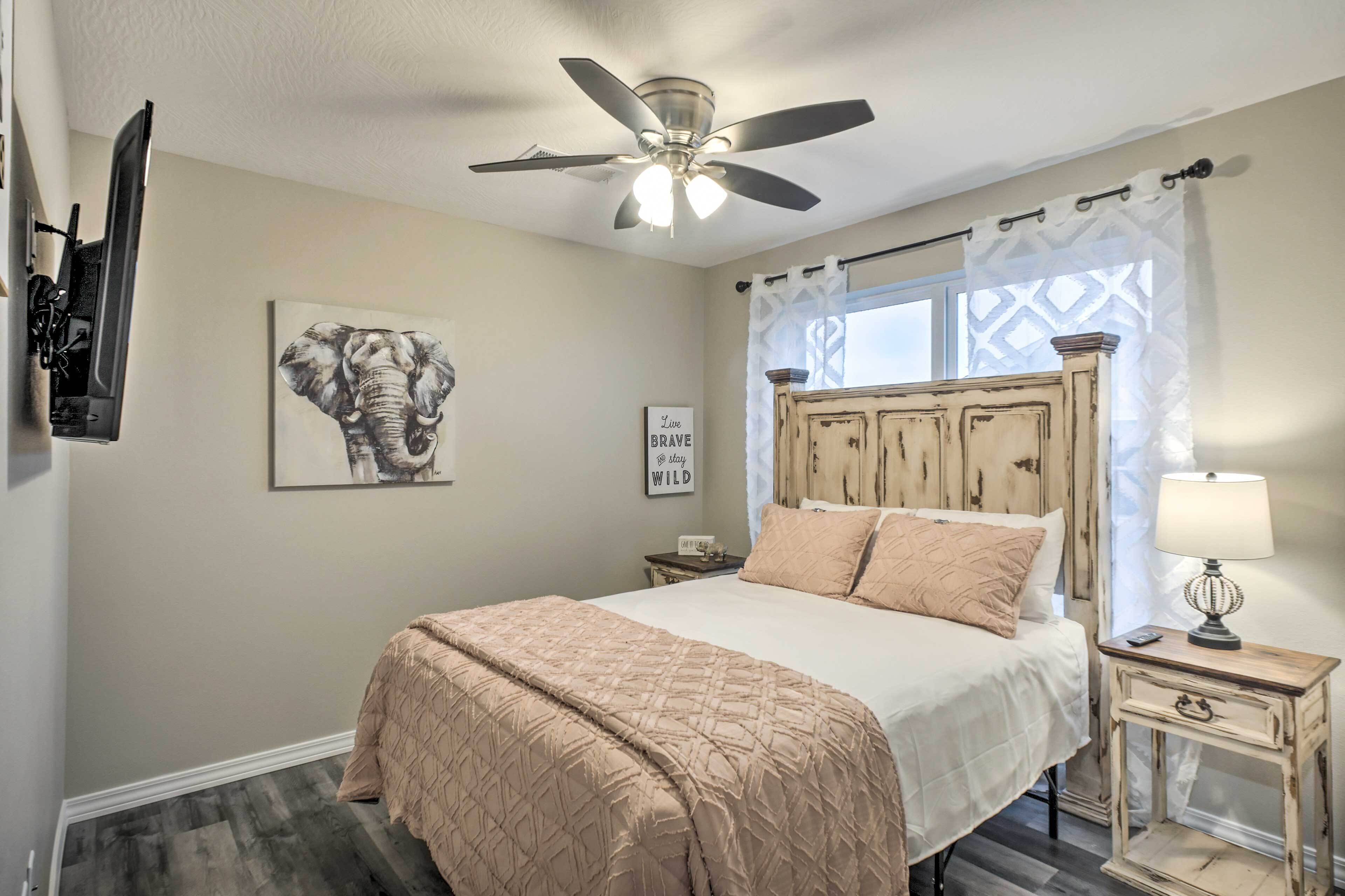 A bedroom with a bed, a ceiling fan, a wall-mounted TV, a window with curtains, and a framed picture.