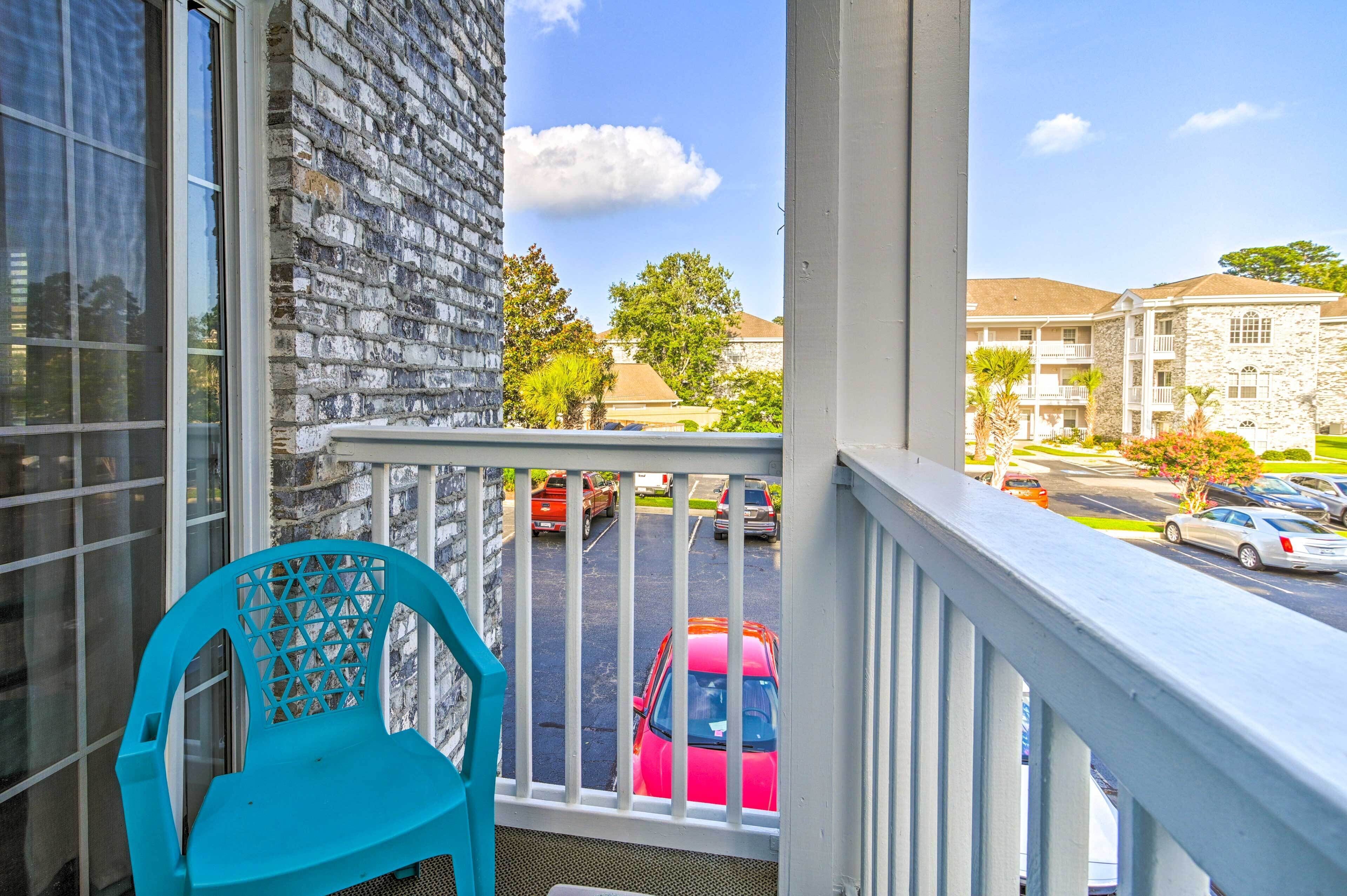 A balcony with a turquoise chair, a stone building, and a parking lot with cars.