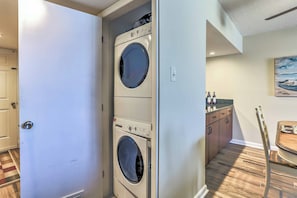 A laundry room with two stacked front-loading washing machines, a wooden floor, a door, and a small kitchenette area with cabinets and a countertop.