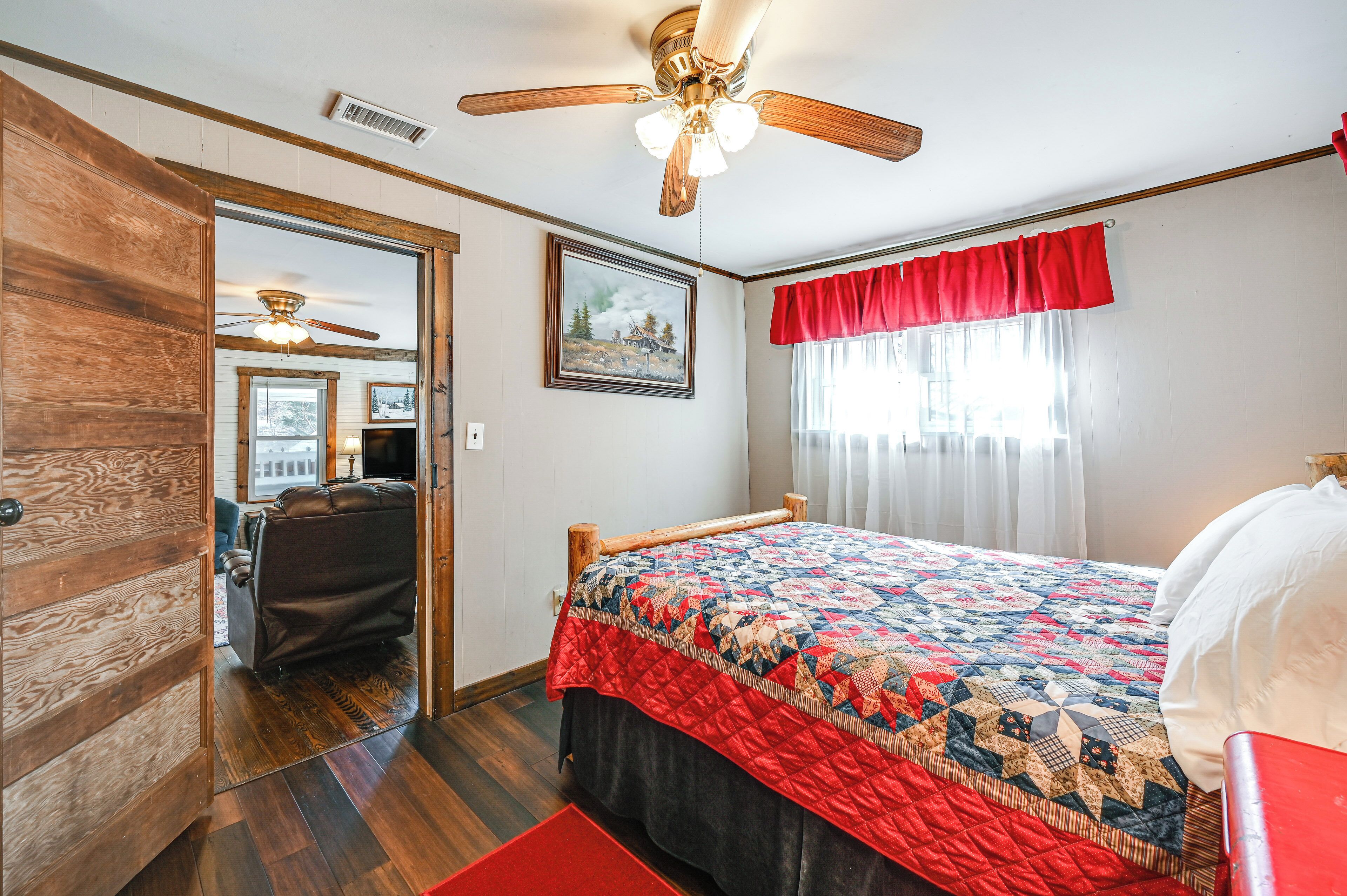 A bedroom with a wooden bed, a ceiling fan, a red window valance, and a painting on the wall.