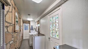 Ein schmaler Korridor mit einer Steinwand auf der einen Seite, einer weißen Tür mit Buntglasfenster auf der anderen und einer kleinen Küchenzeile mit Kühlschrank und Spüle.