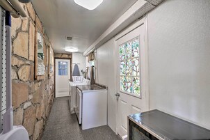Un estrecho pasillo con un muro de piedra en un lado, una puerta blanca con un vitral en el otro, y una pequeña zona de kitchenette con un refrigerador y un lavamanos.