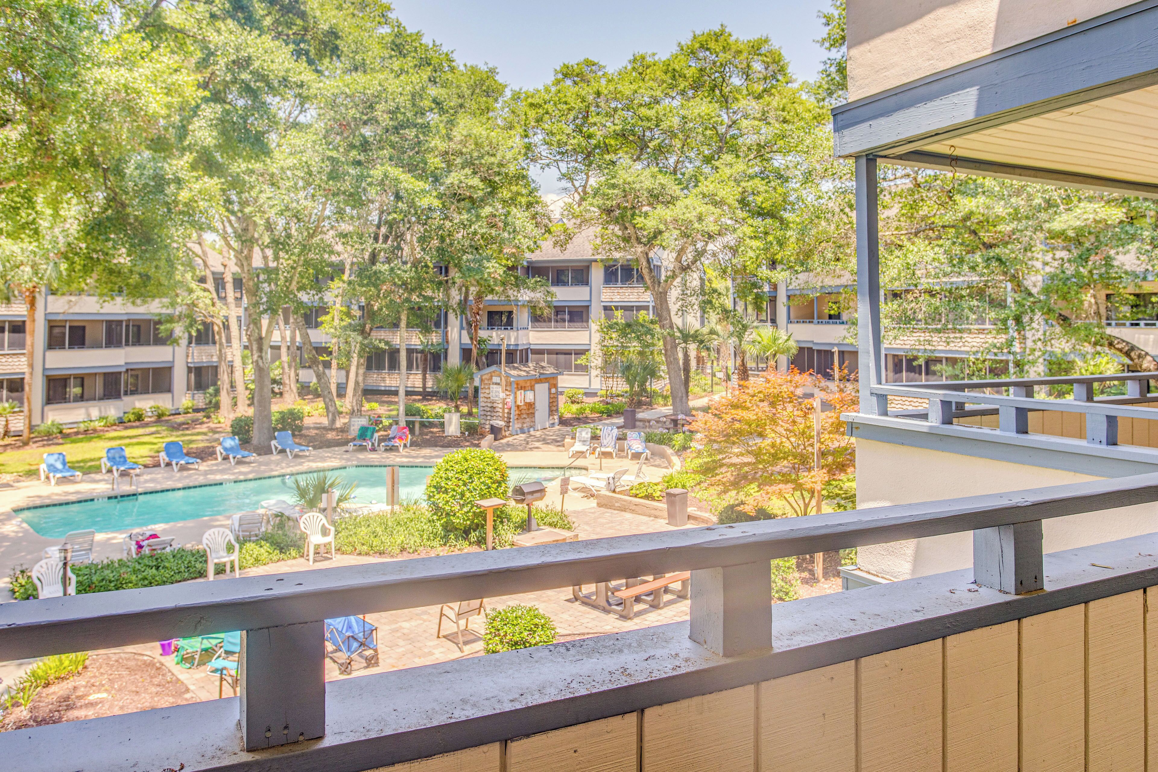 A pool area with lounge chairs and a barbecue grill, surrounded by trees and a building with multiple balconies.