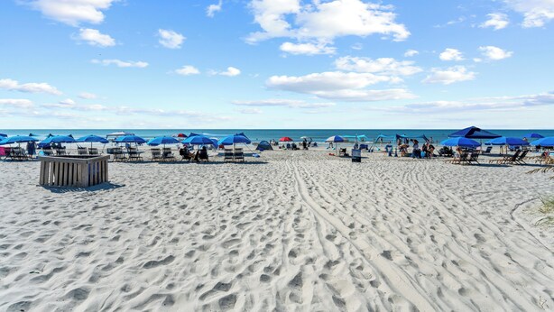 Une plage de sable avec des parasols bleus et des chaises longues, une structure en bois et des empreintes de pas menant à l’eau.