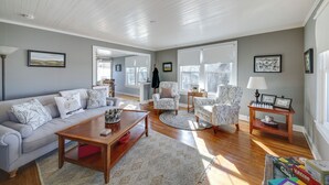 A spacious living room with a light grey sofa, two patterned armchairs, a wooden coffee table, and a round rug. There are large windows allowing natural light, and a small table with a lamp and picture frames. - Cozy Augusta Home w/ Porch: Walk to Katy Trail! (Augusta)