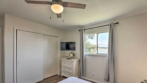 Ein Schlafzimmer mit Bett, Kommode, Wand-Fernseher, Deckenventilator und einem Fenster mit Vorhängen.