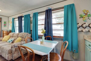 Una sala de estar acogedora con sofá, mesa de comedor y sillas. Hay cortinas en las ventanas y se aprecia un adorno de pájaro en la pared.