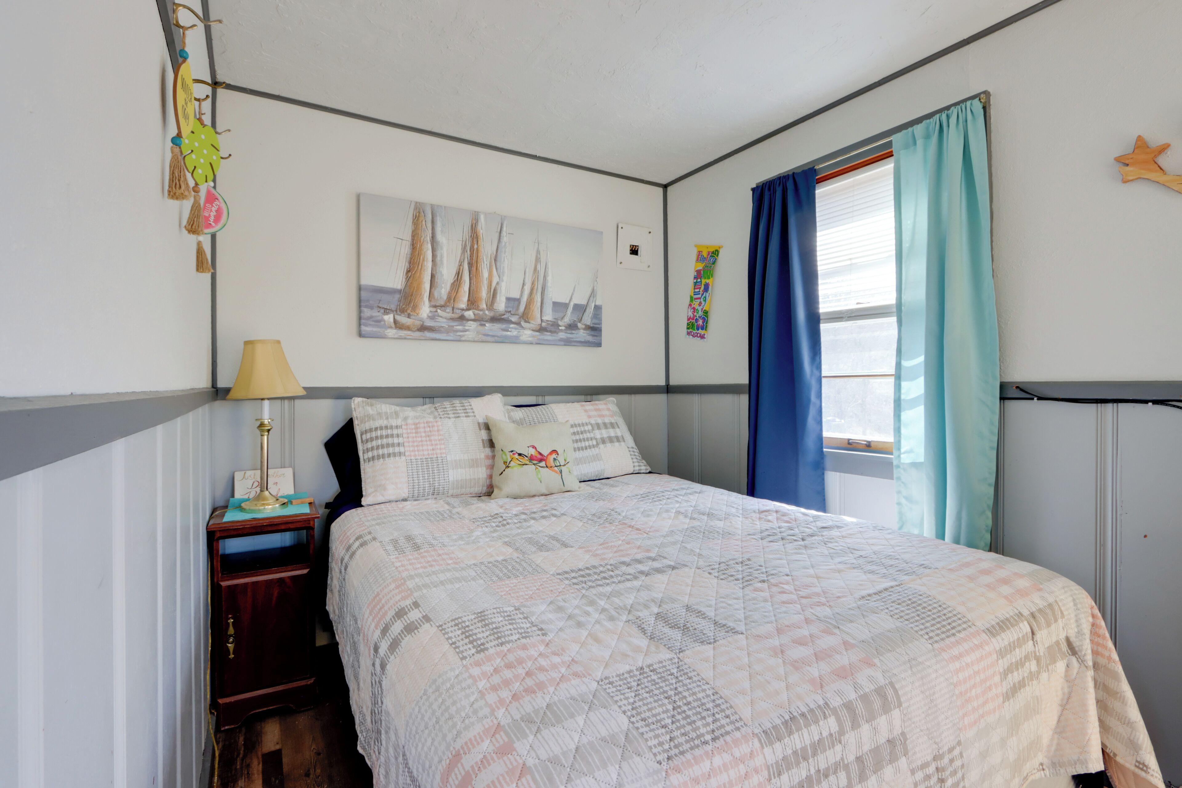 A bedroom with a bed, a nightstand, a lamp, a painting of sailboats, and a window with curtains.