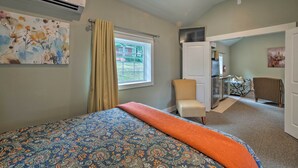Ein Schlafzimmer mit einem Bett, einer Klimaanlage, einem Fenster mit Vorhängen, einem Sessel und einer Tür zu einer Küchenzeile.