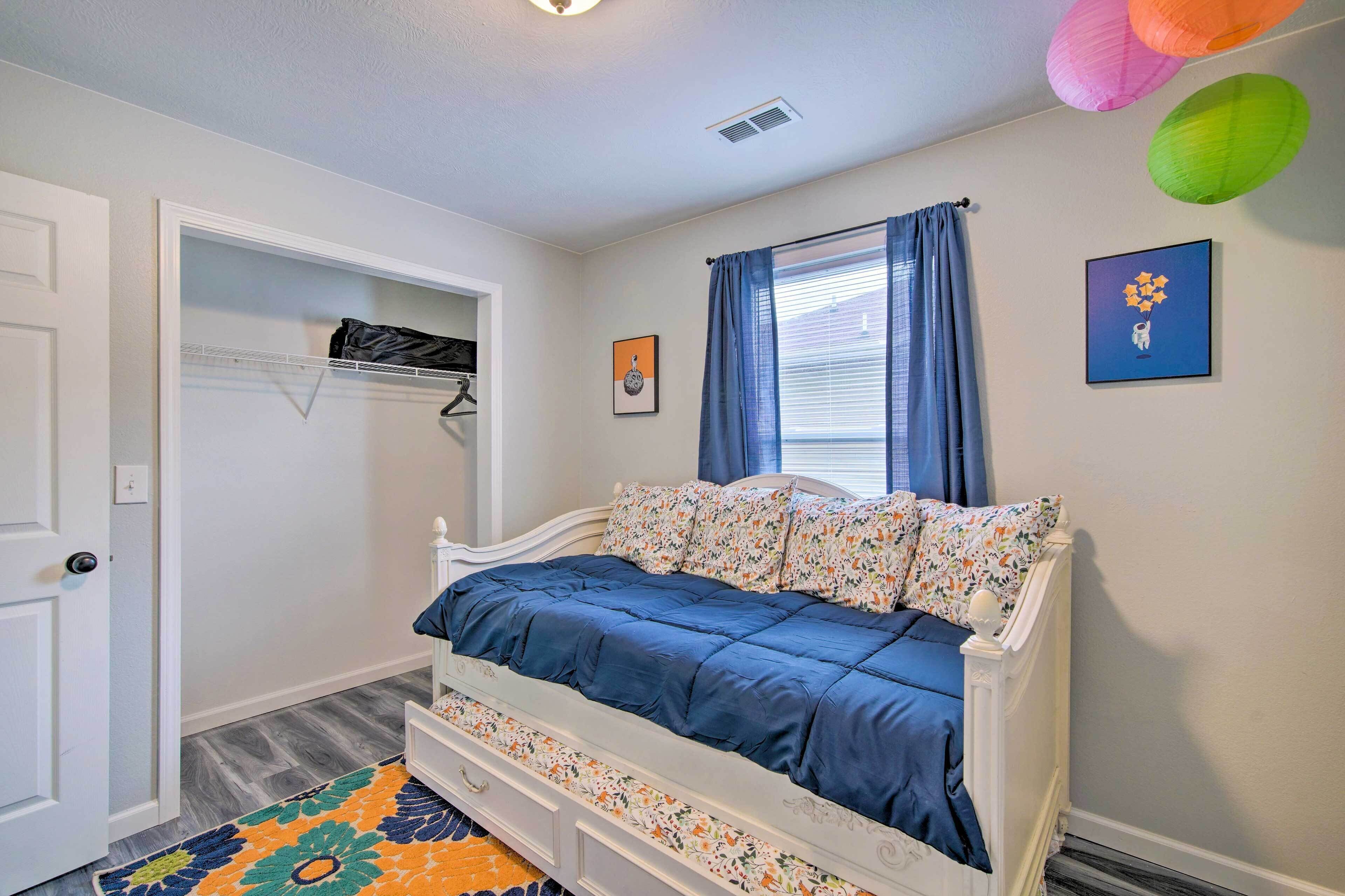 A bedroom with a white bed, floral bedding, a colorful rug, and a window with blue curtains.