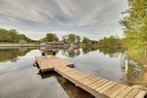A wooden dock extending into a calm lake with a reflection of buildings and trees. - Waterfront Home Near Mark Twain National Forest (Poplar Bluff)