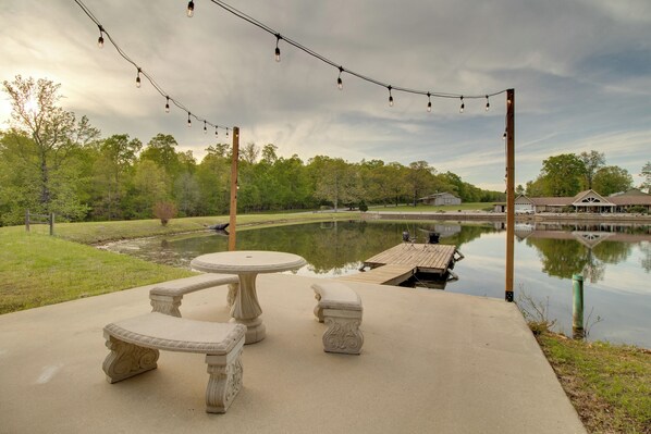 A concrete patio with a table and benches overlooking a calm pond, surrounded by trees and a wooden dock. - Waterfront Home Near Mark Twain National Forest (Poplar Bluff)