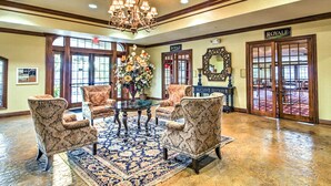 Um hall com um candelabro, poltronas estampadas, uma mesa central com um vaso de flores e uma placa com a inscrição "ROVALE".