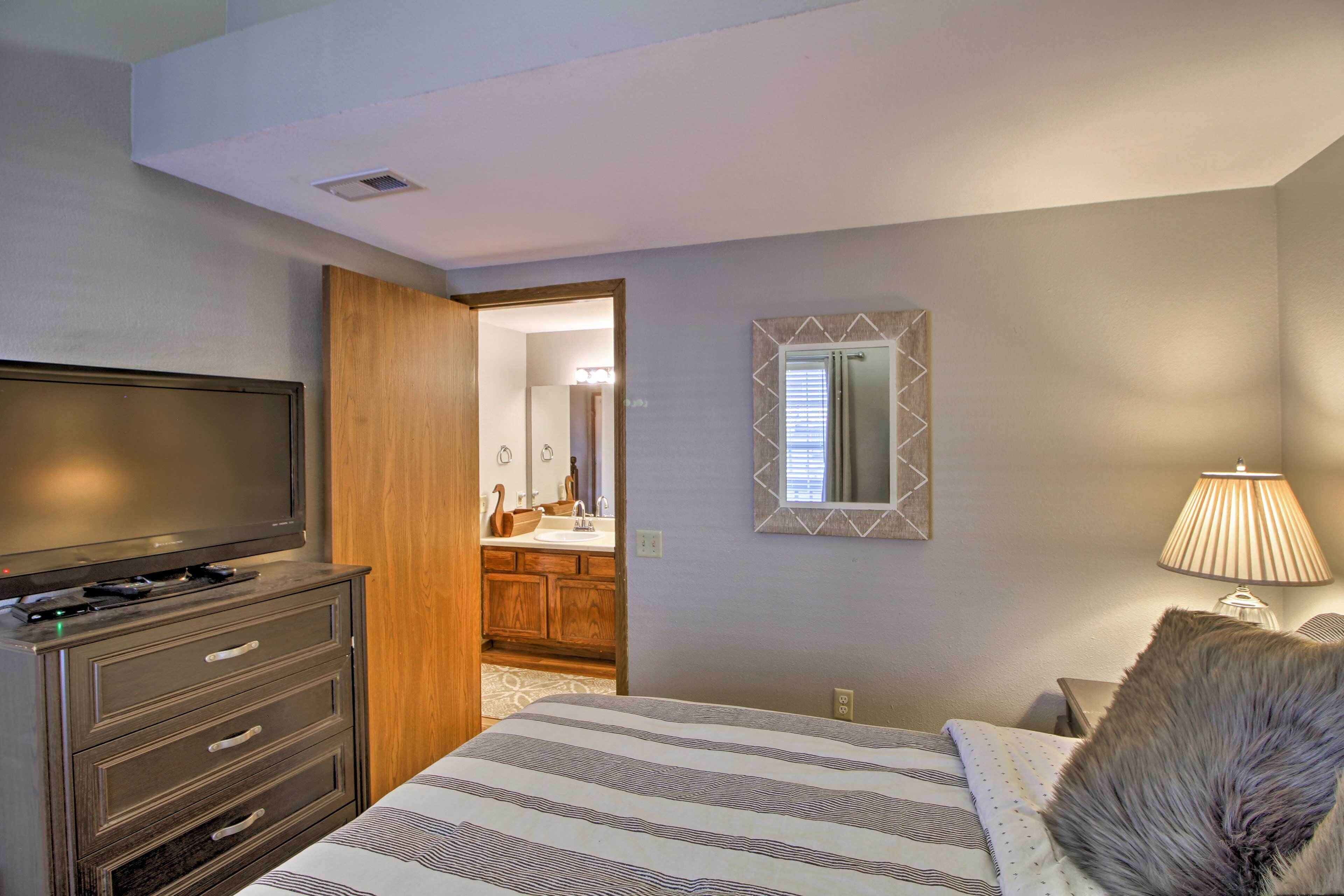 Un dormitorio con cama, cómoda, televisor, espejo y un baño visible a través de una puerta abierta.