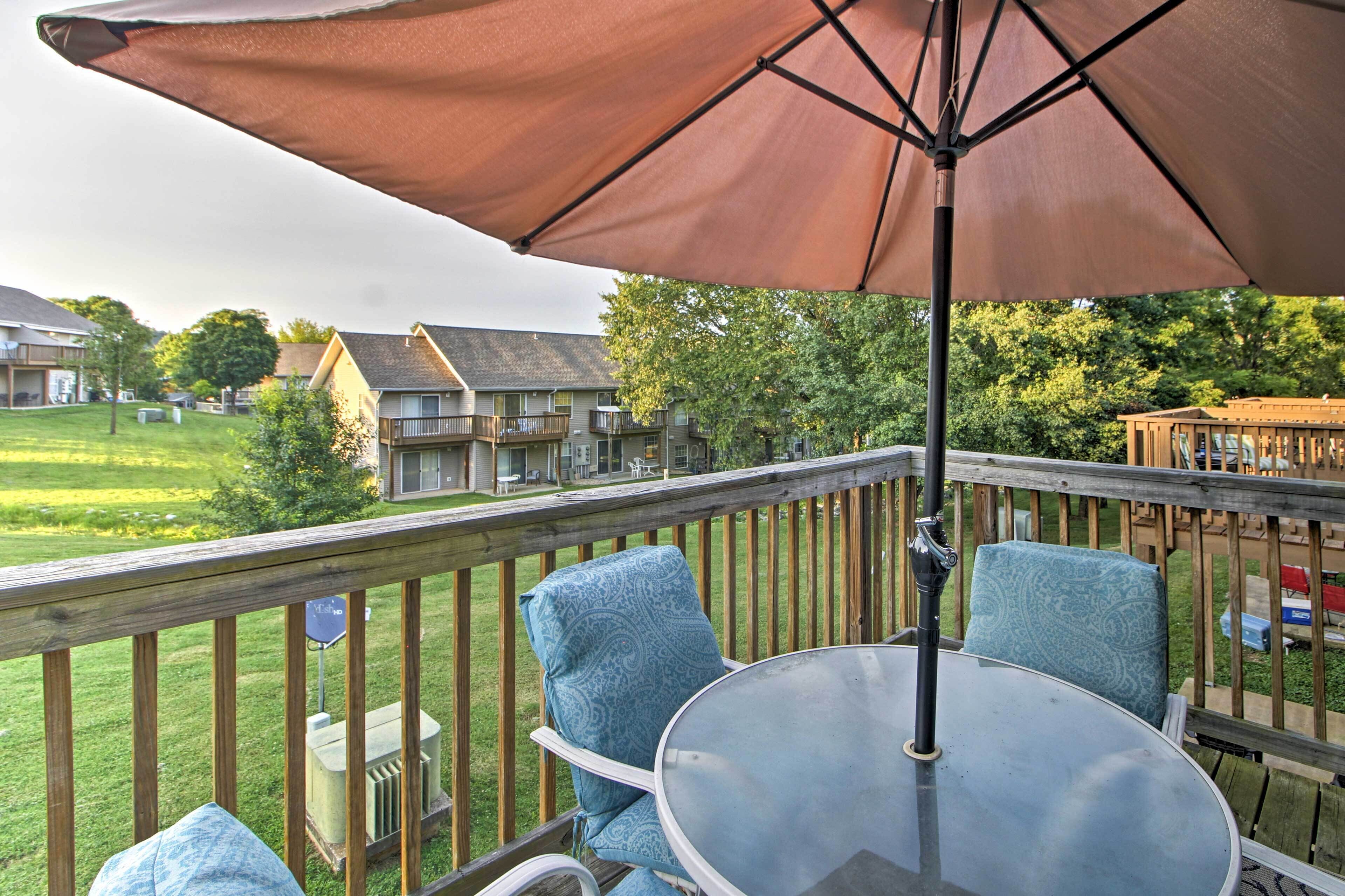 A deck with a glass table and blue chairs, overlooking a grassy area and residential buildings.