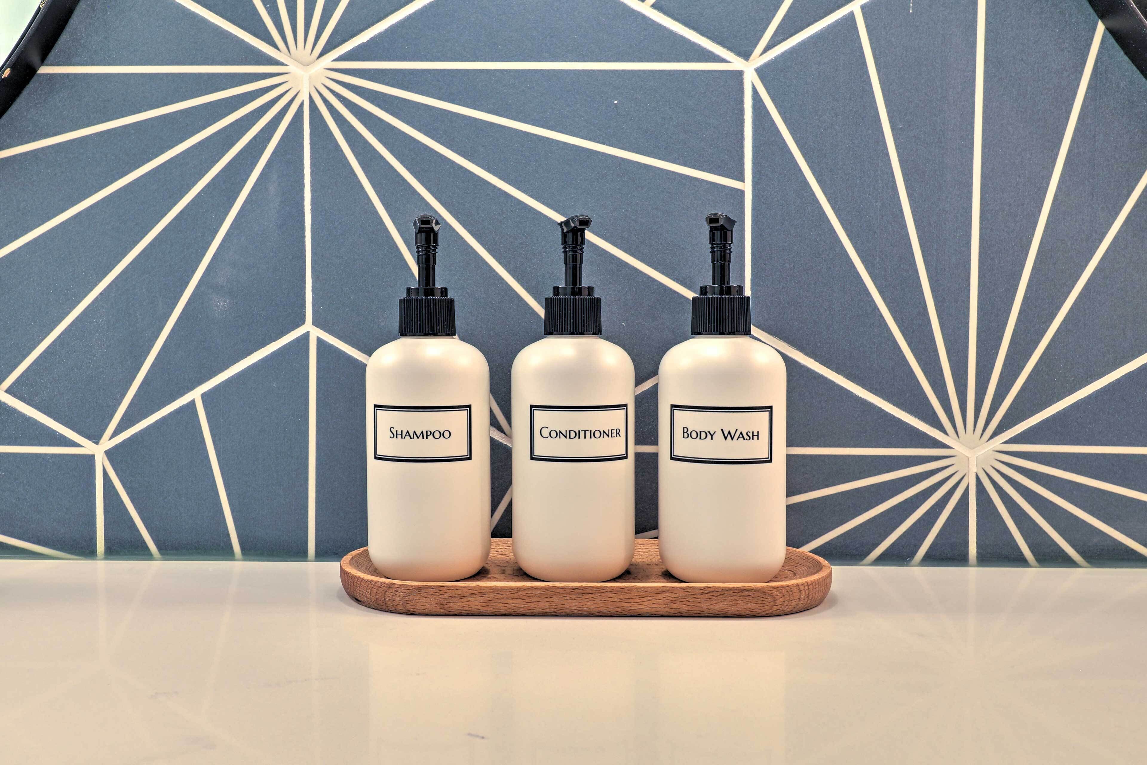 Three bottles of shampoo, conditioner, and body wash on a wooden tray against a geometric-patterned wall.