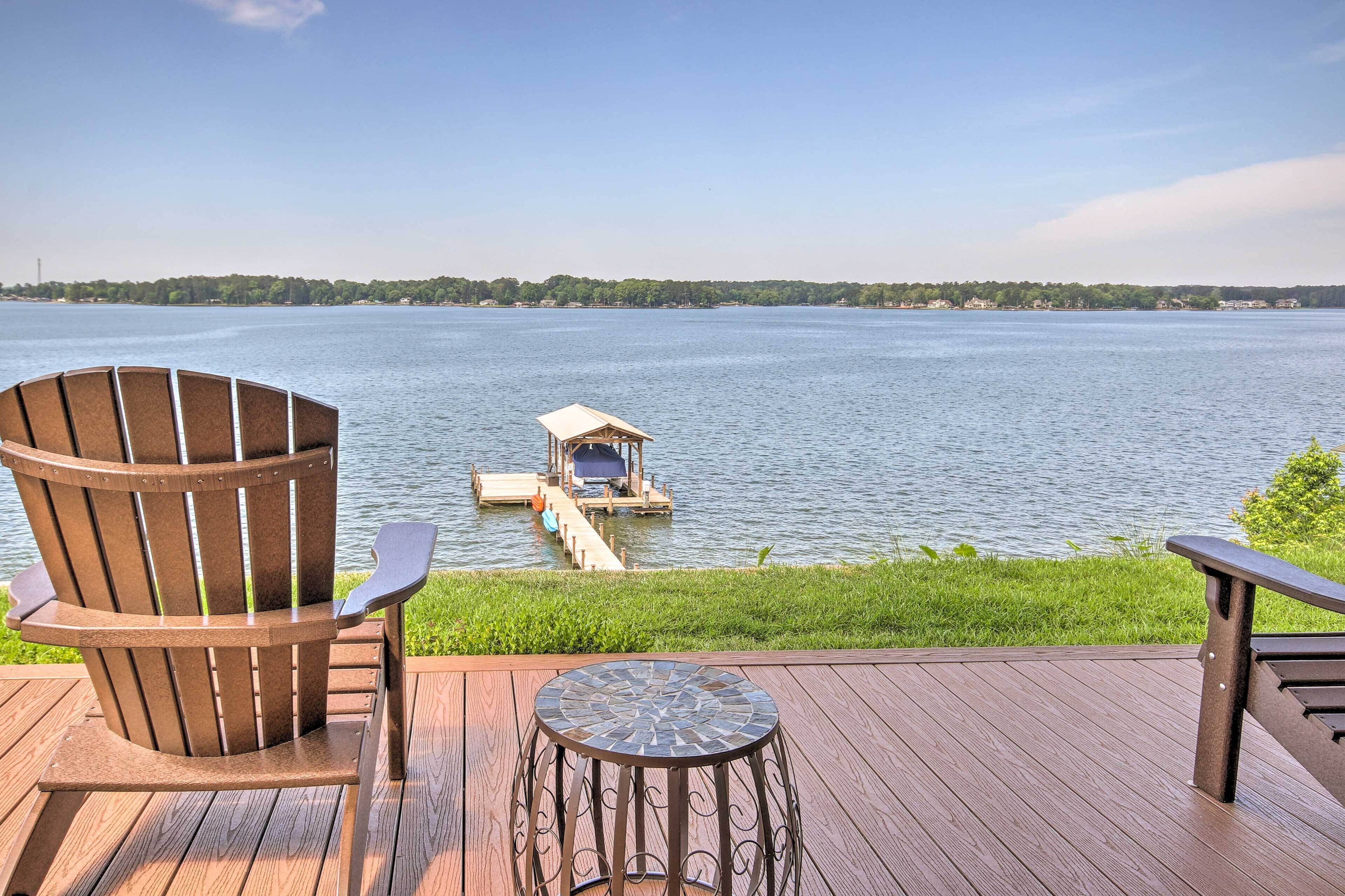 A wooden deck with a Adirondack chair and a small table, overlooking a calm lake with a dock and a small structure.