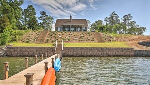 Une maison au bord du lac, avec une jetée qui s’avance dans l’eau, un mur de soutènement en pierre et une colline herbeuse avec des marches menant à la maison.