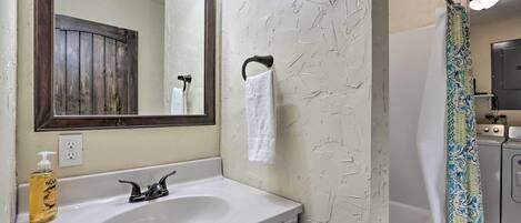A bathroom with a white sink, a mirror, a towel hanging on a hook, a shower curtain, and a washing machine.