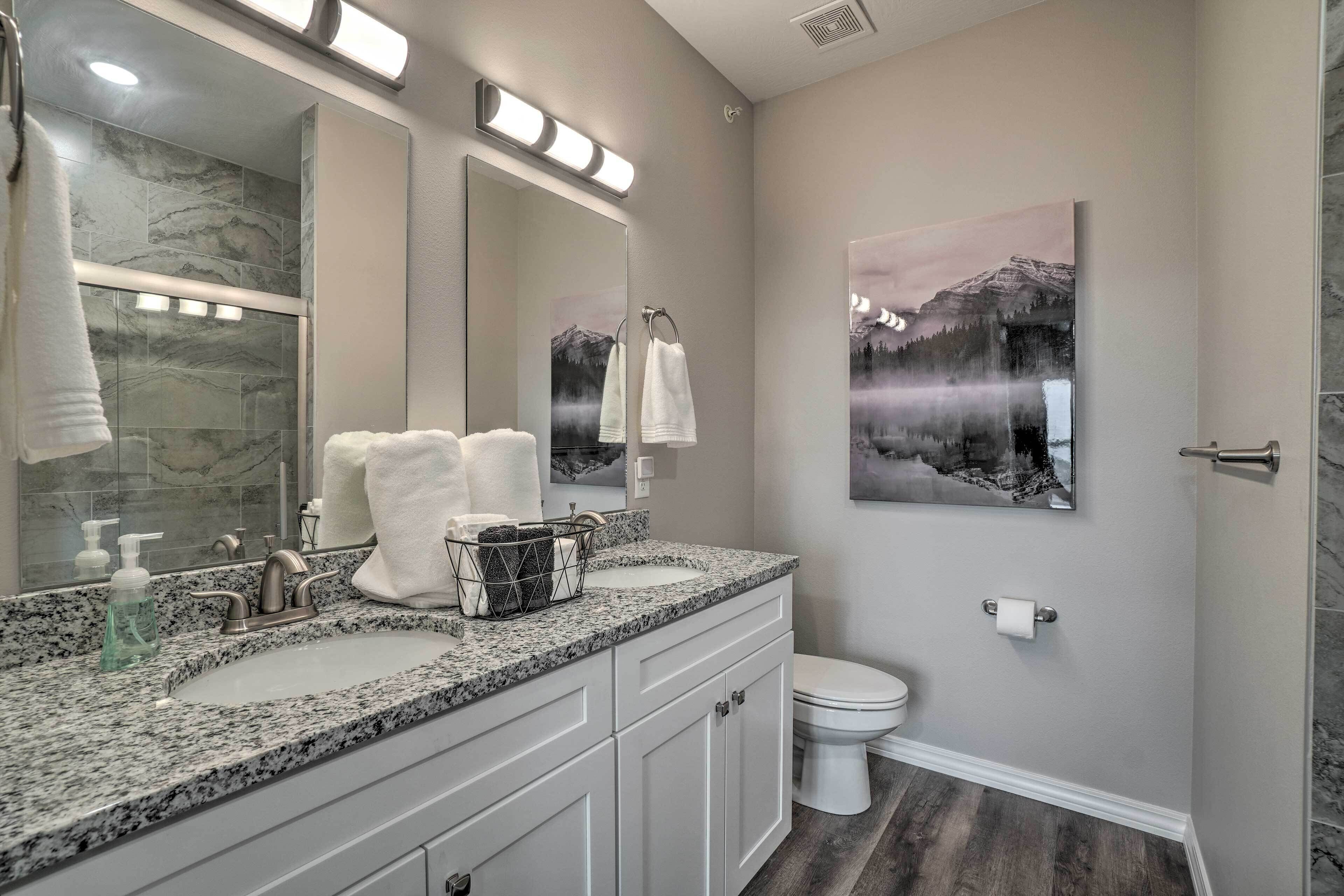 A bathroom with a granite countertop, white cabinets, a large mirror, a sink, a toilet, and a framed picture on the wall.
