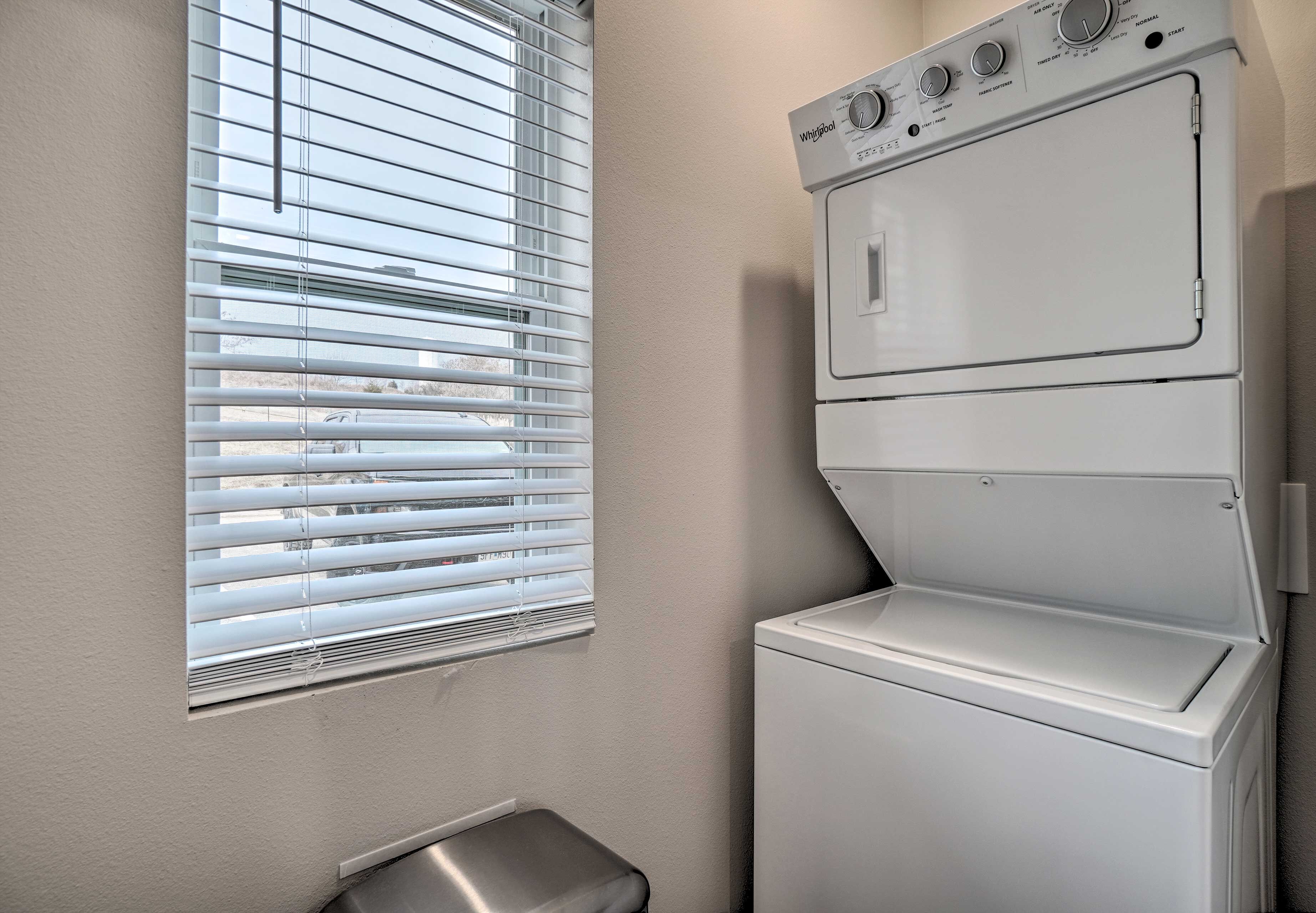 A laundry area with a stacked washer and dryer, a window with blinds, and a small bench.