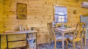 A wooden cabin interior with a dining table, chairs, a small desk with a radio, and a window with curtains.