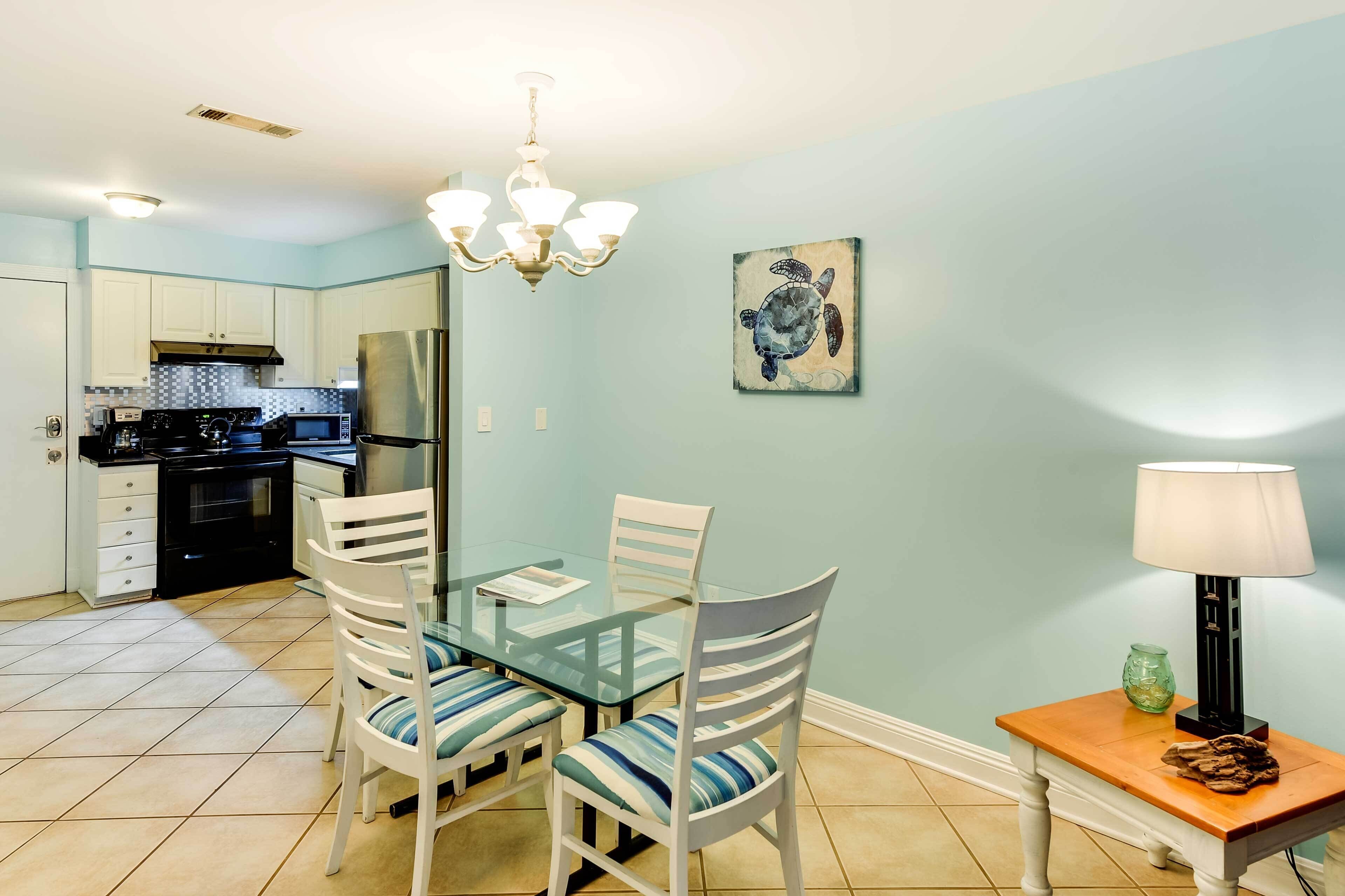 A dining area with a glass table and white chairs, a kitchen in the background, and a turtle painting on the wall.