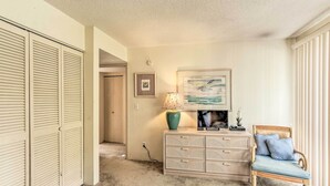 A room with a beige carpet, a wooden dresser, a wicker chair, a lamp, and a framed picture on the wall. - Patio & Walk to Beach: Hilton Head Island Townhome (Hilton Head Island)