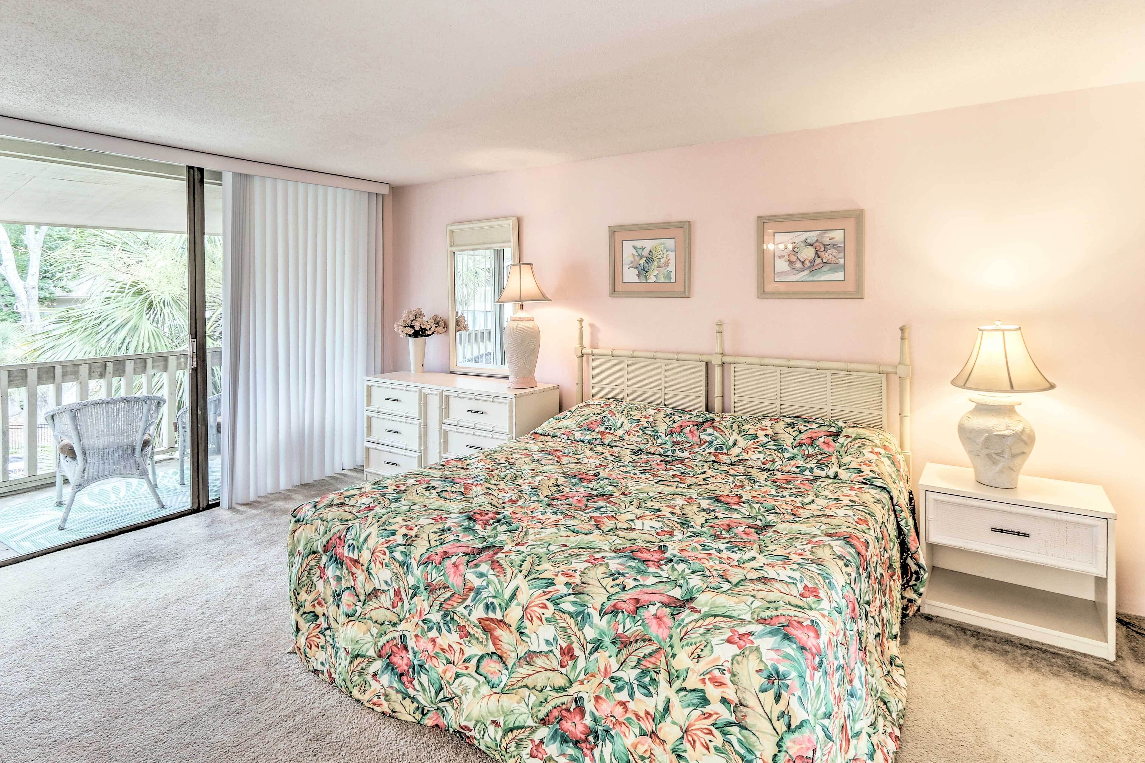 A bedroom with a floral bedspread, a white nightstand with a lamp, a white dresser, and a sliding glass door leading to a balcony.