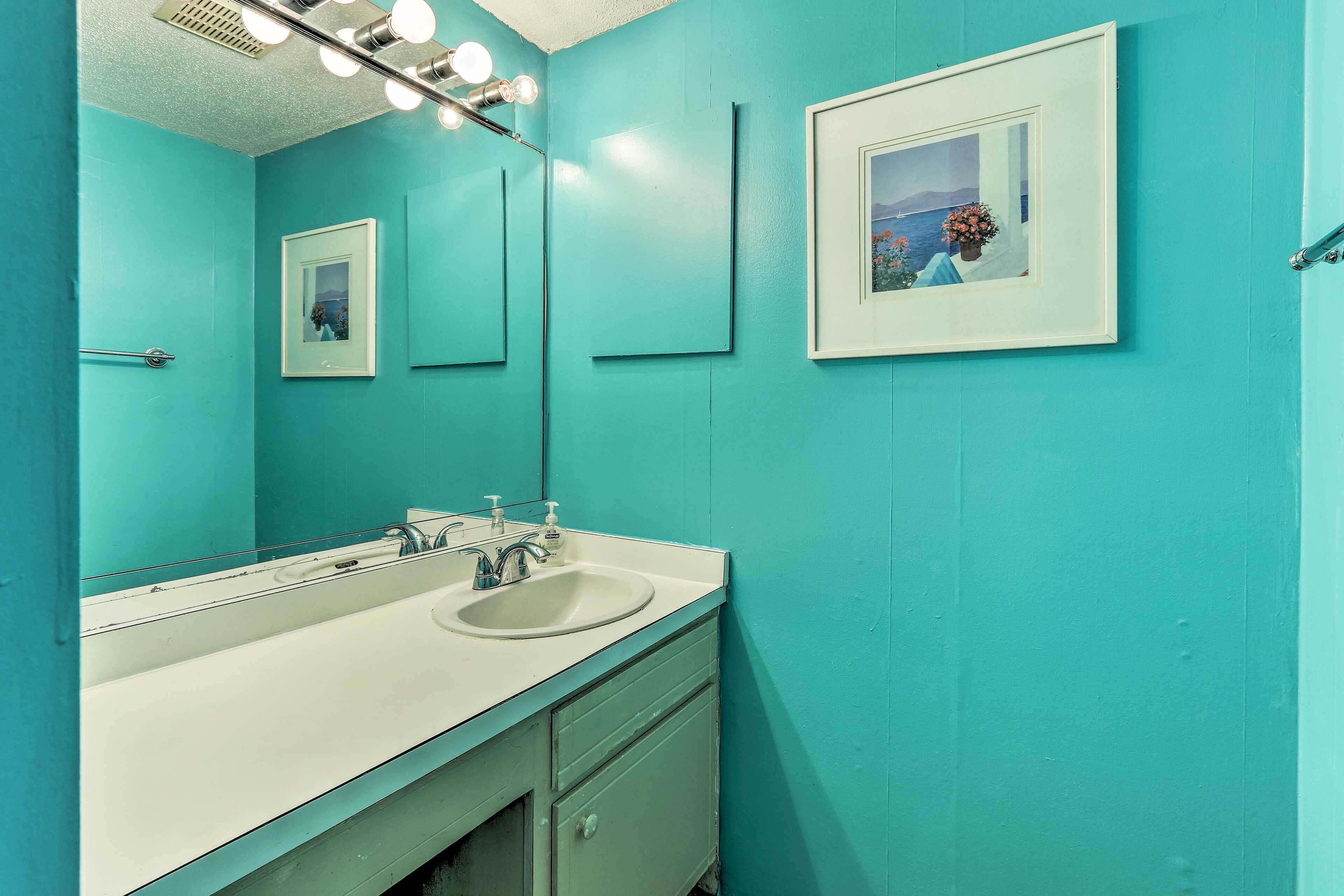 A bathroom with a turquoise wall, a white sink, and a mirror with lights above it. There are framed pictures on the wall.