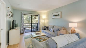 A living room with a sofa, a coffee table, and a television on a stand. There is a window with a view of trees and a door leading to an outdoor area.