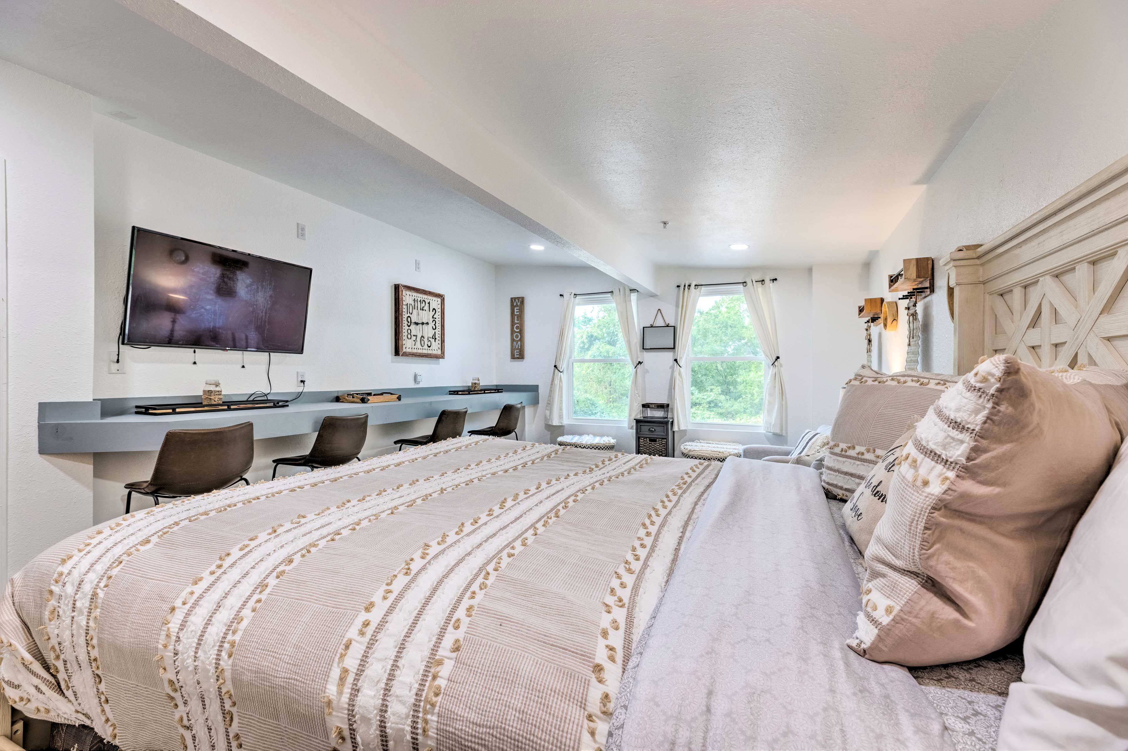 A bedroom with a large bed, a TV mounted on the wall, a desk with chairs, and a window with curtains.