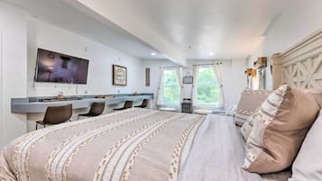 A bedroom with a large bed, a TV mounted on the wall, a desk with chairs, and a window with curtains.