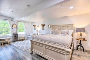 Quarto com cama grande, mesa de cabeceira, cadeira, mesa redonda pequena e janela com cortinas.