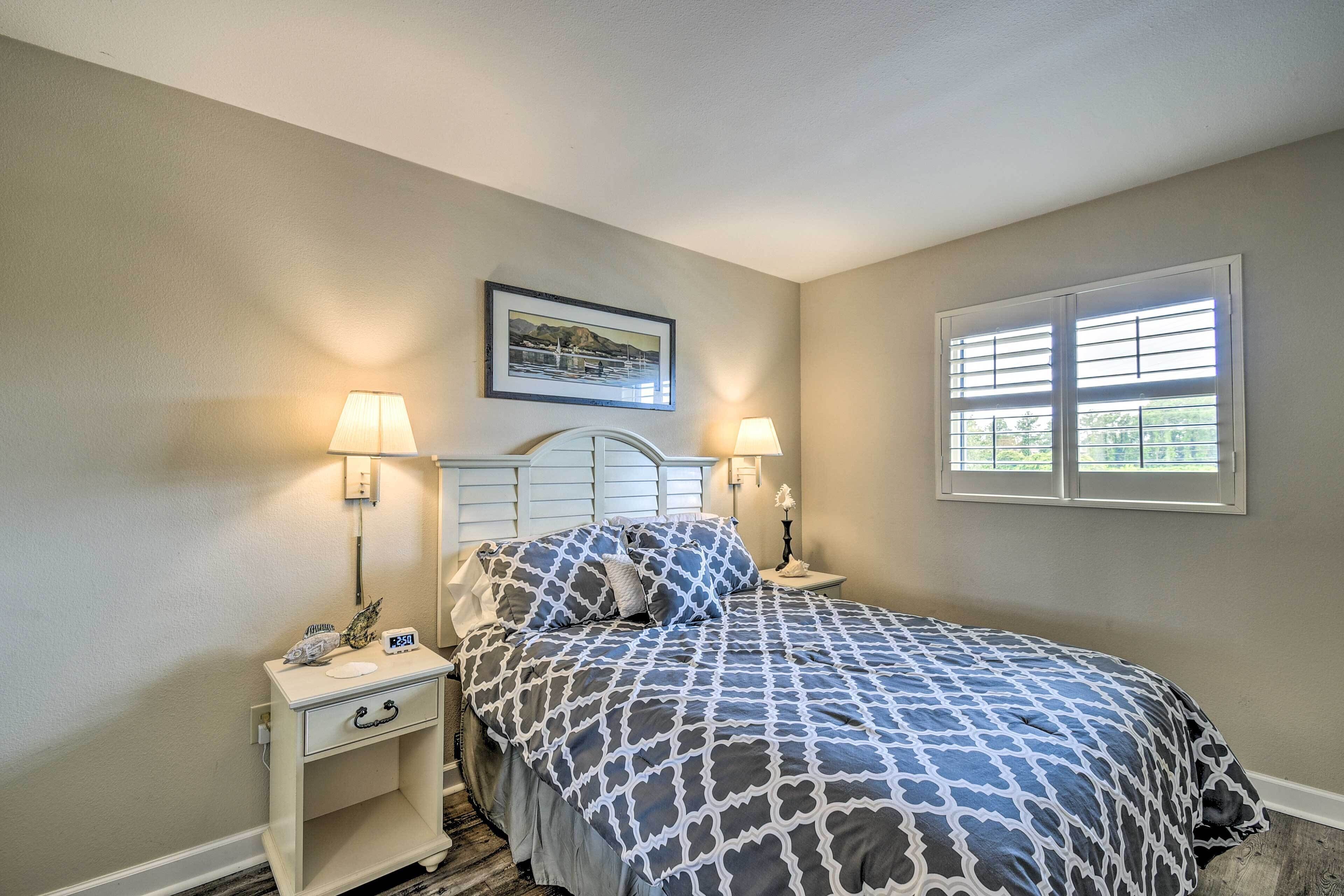A bedroom with a bed, two bedside tables, a nightstand, a framed picture on the wall, and a window with blinds.