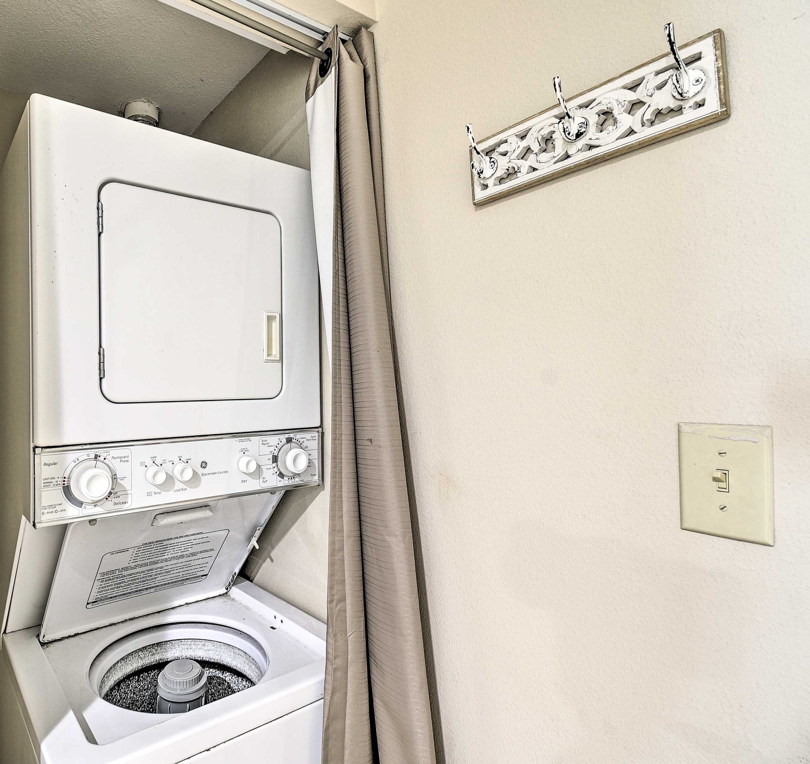 A compact, white washing machine with multiple control knobs and a front-loading door, situated in a small laundry room with a beige wall and a curtain partially drawn.