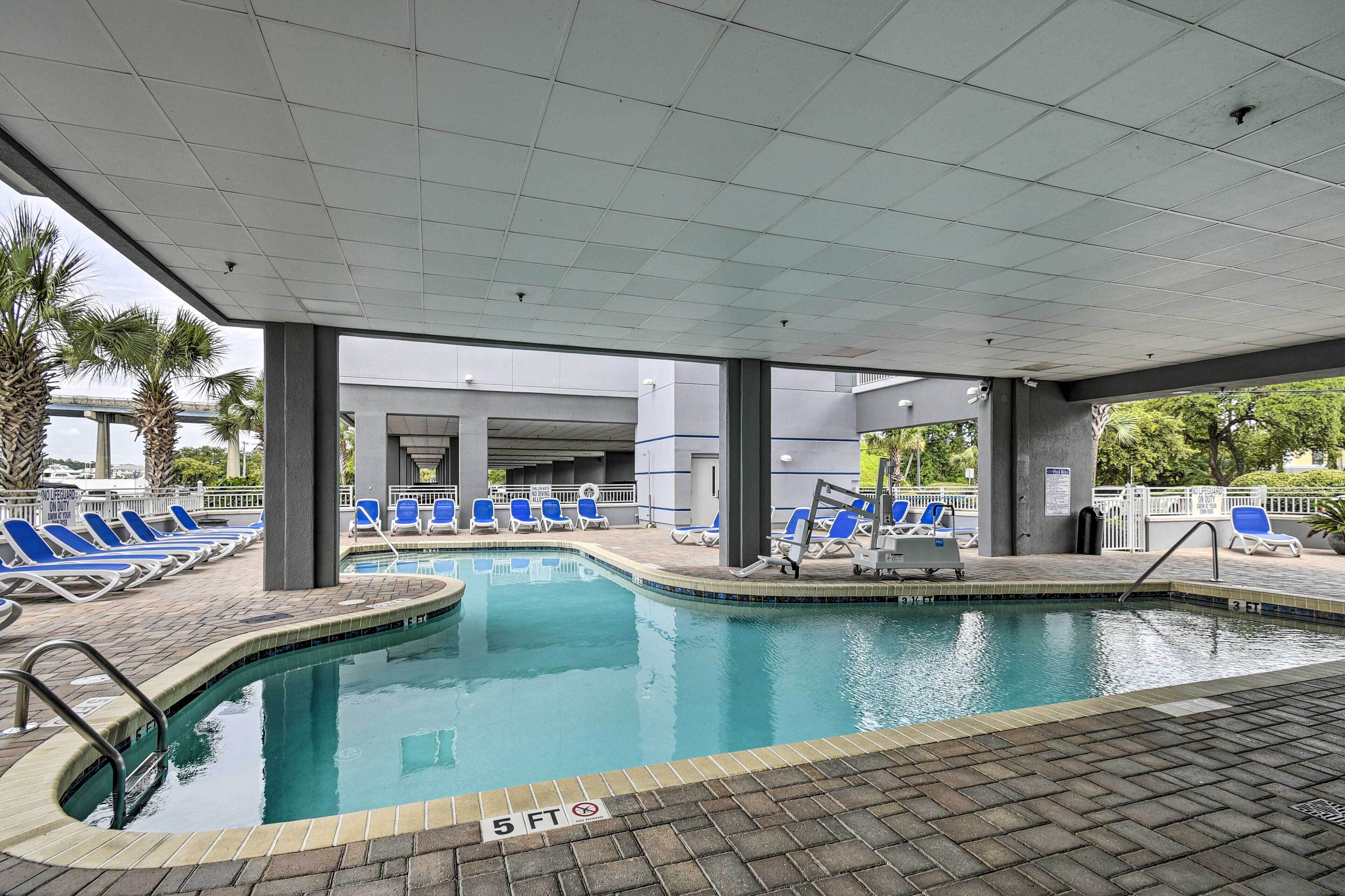 A swimming pool with blue lounge chairs, a tiled deck, and a covered area with pillars.