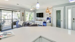 A modern living room with a white sofa, blue armchair, and a large white coffee table. There is a TV mounted on the wall and a ceiling fan. - Walk to Coligny Beach: Tropical Hilton Head Condo (Hilton Head Island)