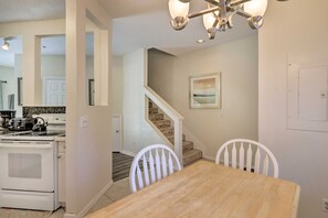 Un comedor con una mesa de madera y sillas blancas, una cocina con electrodomésticos blancos y una escalera que sube a otro nivel.