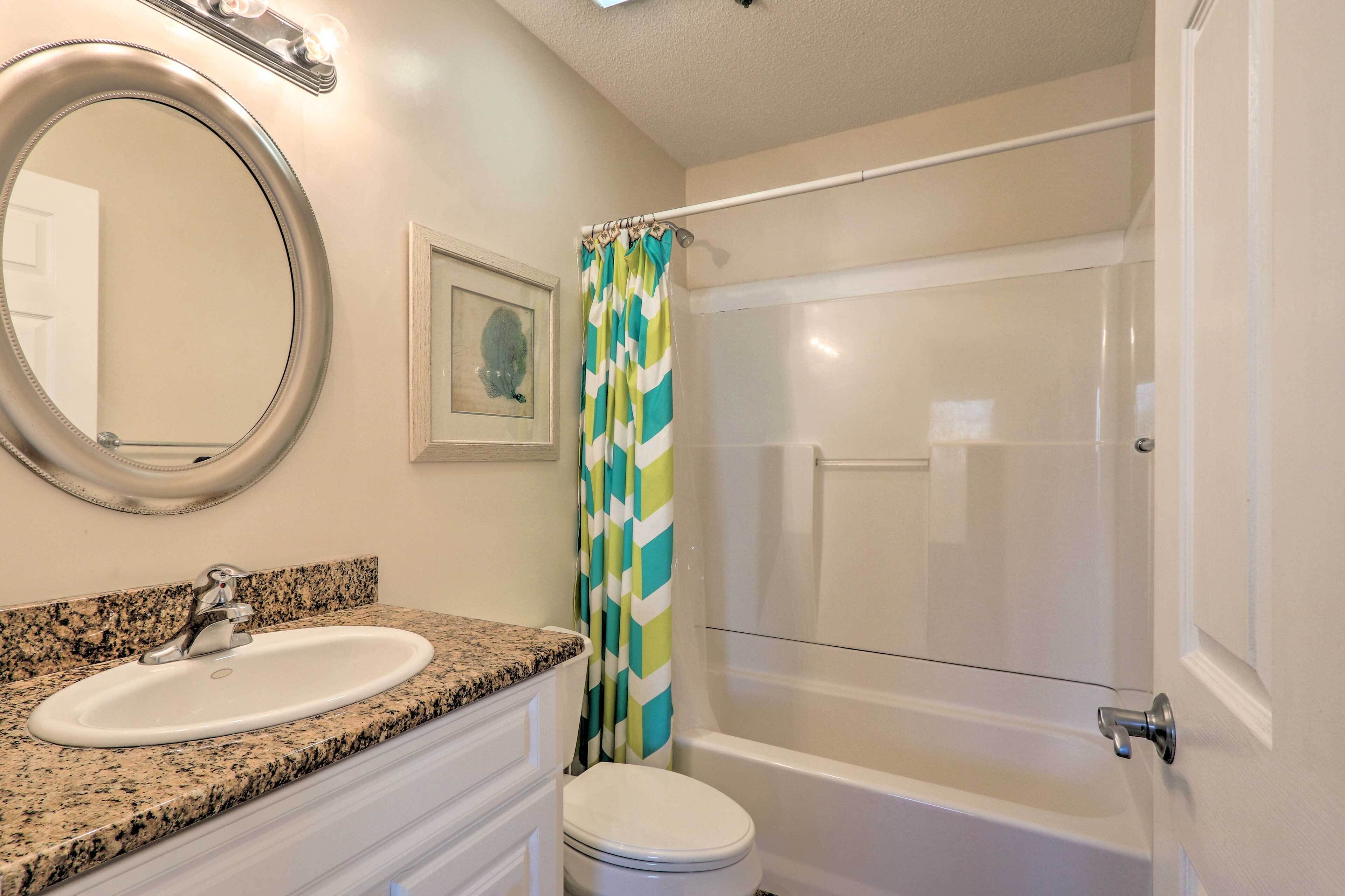 Une salle de bain avec un lavabo surmonté d’un plan de travail en granit, des toilettes blanches, une douche dotée d’un rideau à rayures vertes et blanches, et un miroir avec éclairage intégré.