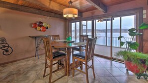 A dining area with a wooden table and chairs, a hanging light fixture, and a view of a lake through large windows. - Inviting Home w/ Pool & Hot Tub on Table Rock Lake (Lampe)