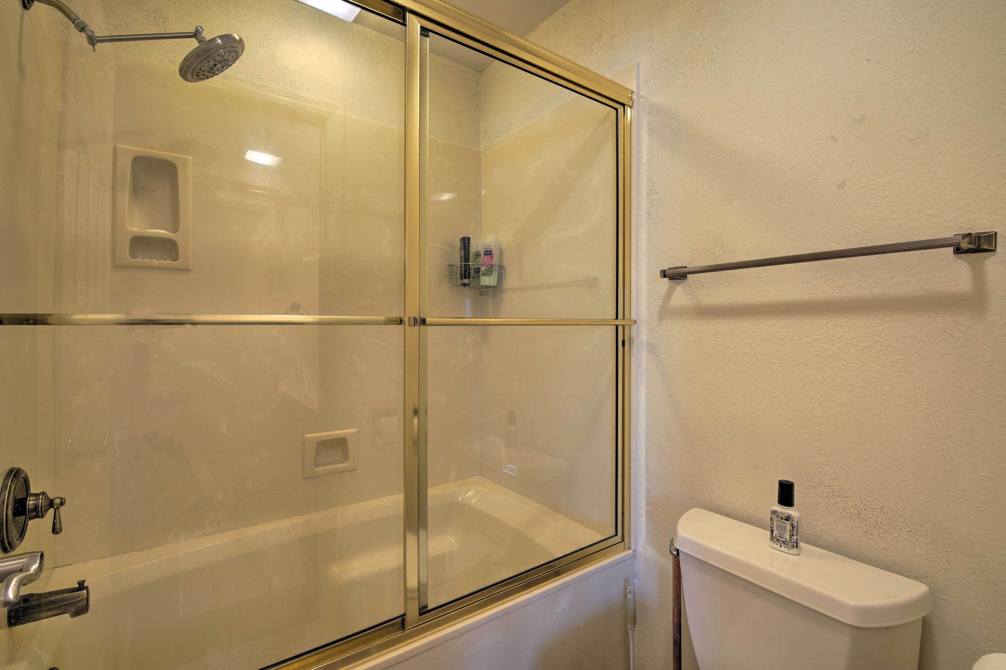 A glass-enclosed shower area with a handheld showerhead, a glass door, and a toilet with a bottle of hand soap on the tank.