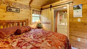 A wooden cabin bedroom with a bed, a nightstand, a lamp, a window with curtains, a calendar, and a door leading to a bathroom. - Creek Views & Grill: Idyllic Florence Cabin (Florence)