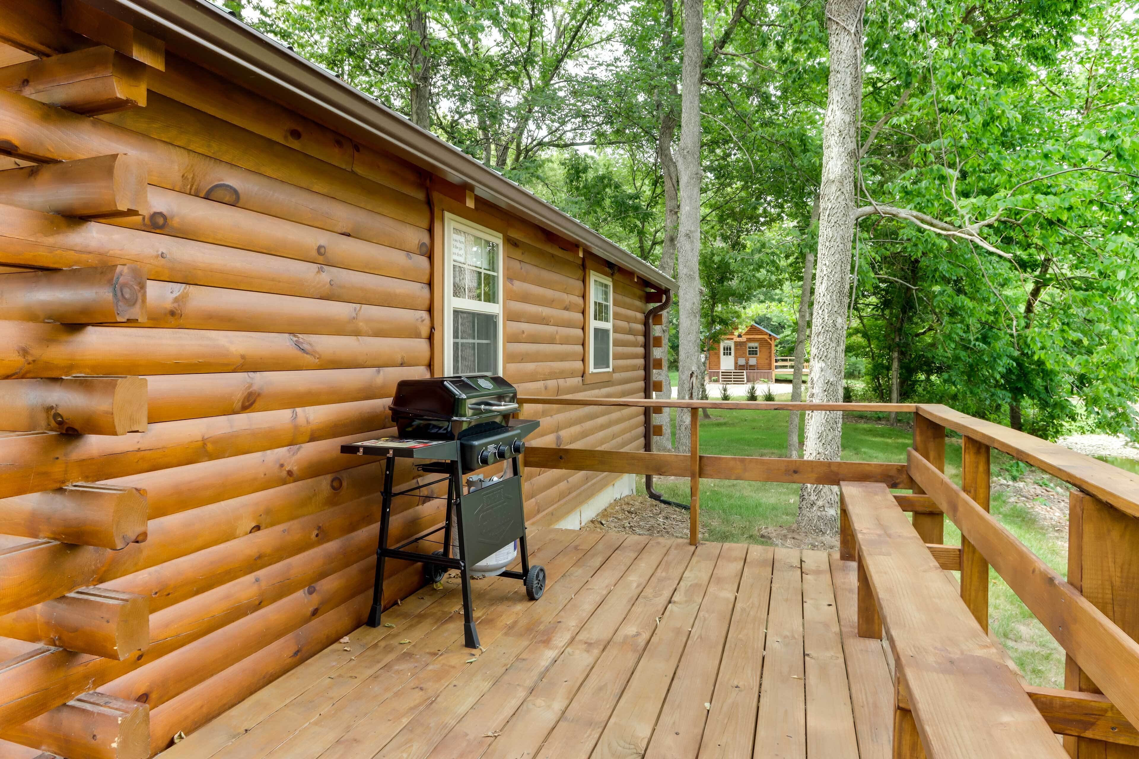 Une terrasse en bois avec un barbecue et une poubelle, entourée d’arbres.