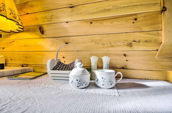 Interior de una cabaña de madera con una mesa dispuesta con teteras y tazas blancas, un cuaderno y un cojín a cuadros.