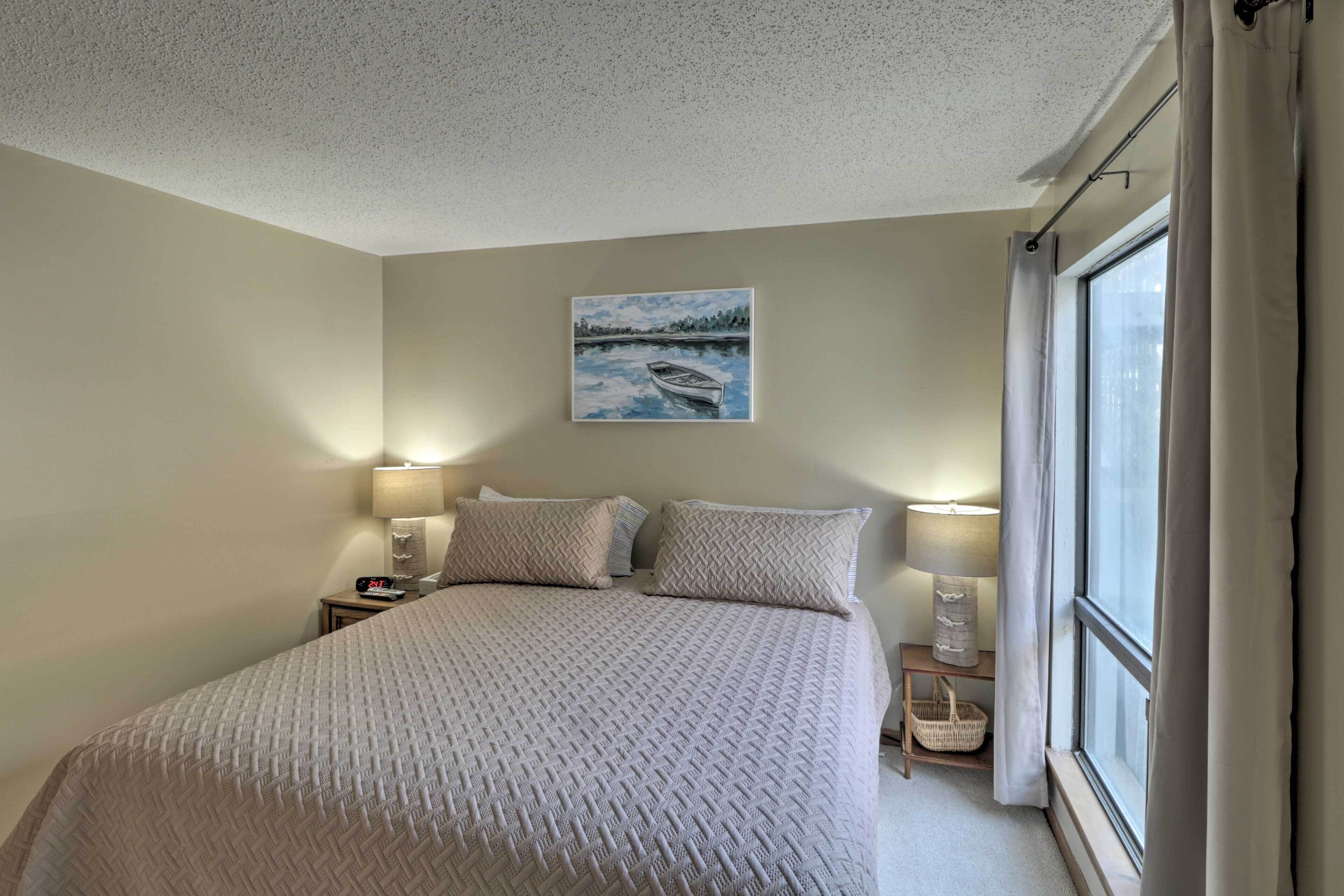 A neatly made bed with a textured blanket, two bedside lamps, a small bedside table with a lamp, a painting of a lake scene, and a window with curtains.