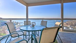 Un espace extérieur aménagé, abrité et doté d’une table et de chaises en verre, offrant une vue sur une étendue d’eau et sur le paysage urbain au loin.