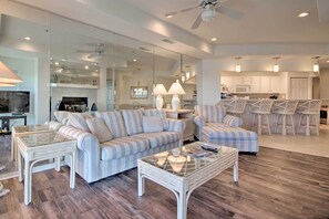 A spacious living room with a striped sofa, a glass coffee table, and a dining area with bar stools. - Pool + Water Views! 3rd-floor Lake Ozark Retreat (Lake Ozark)