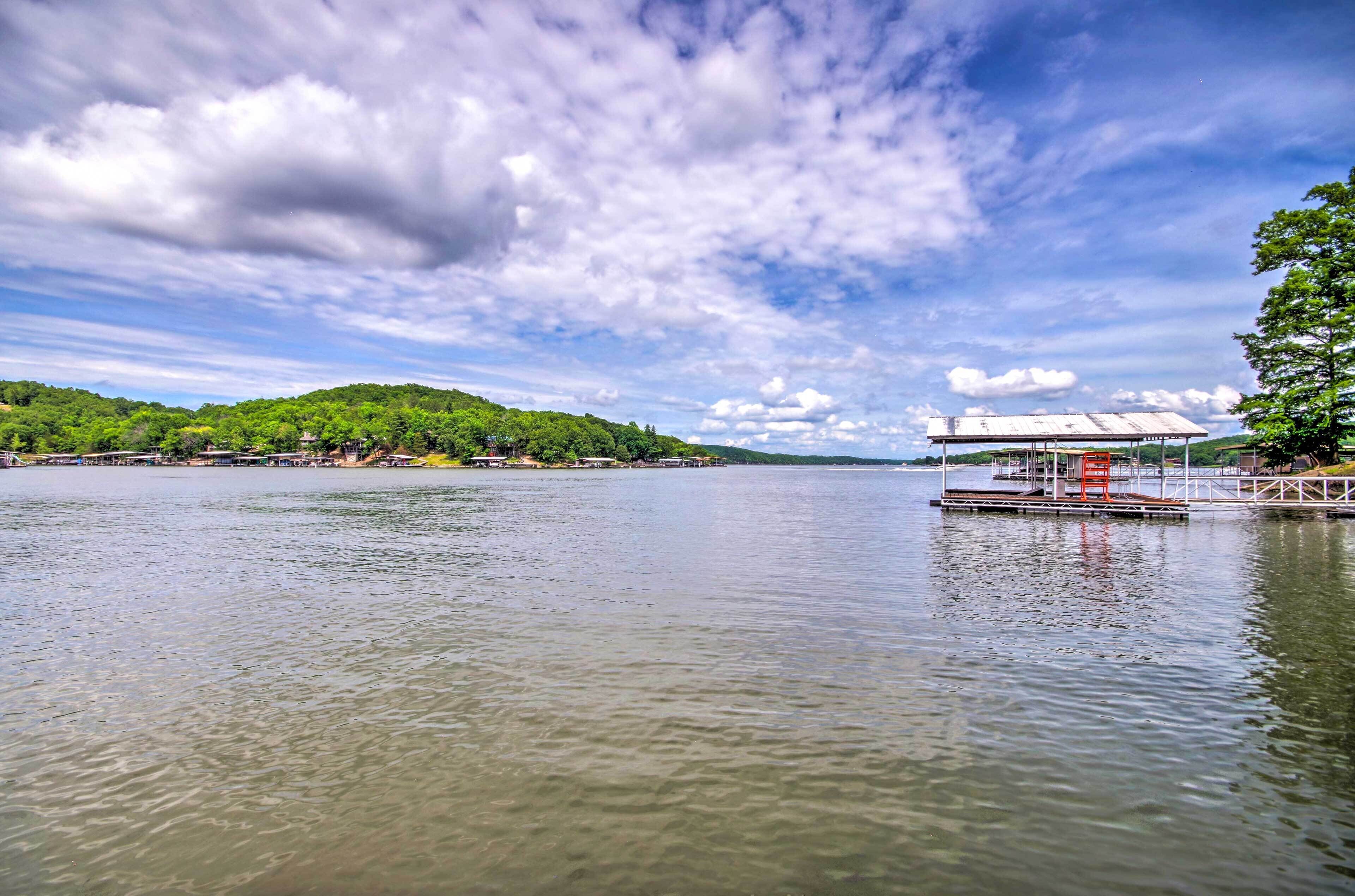 A calm lake with a dock and a covered area, surrounded by greenery and a clear sky.